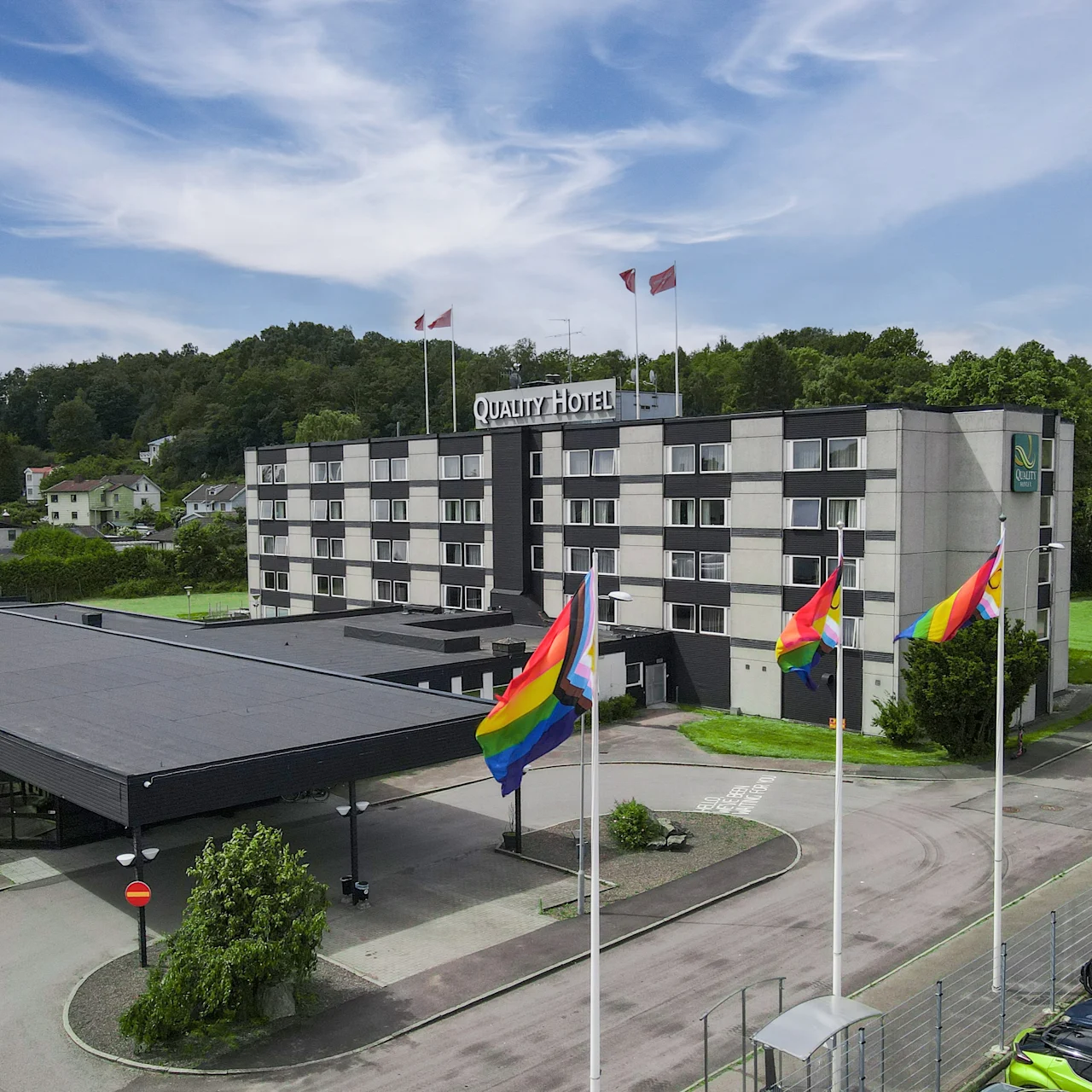 Et fleretagers hotel har regnbueflag på indgangen, omgivet af træer og bakker. Tekst: "Quality Hotel."