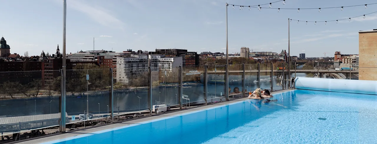 Et basseng på taket med to personer som slapper av ved kanten, omgitt av lysslynger. Utsikt over en bylandskap med bygninger og en elv under en klar himmel.