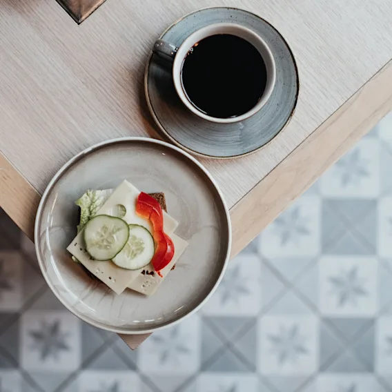 En smörgås med ost, gurka och paprika på en tallrik bredvid en kaffekopp på ett ljust träbord. Golvet har blåvita mönstrade plattor.