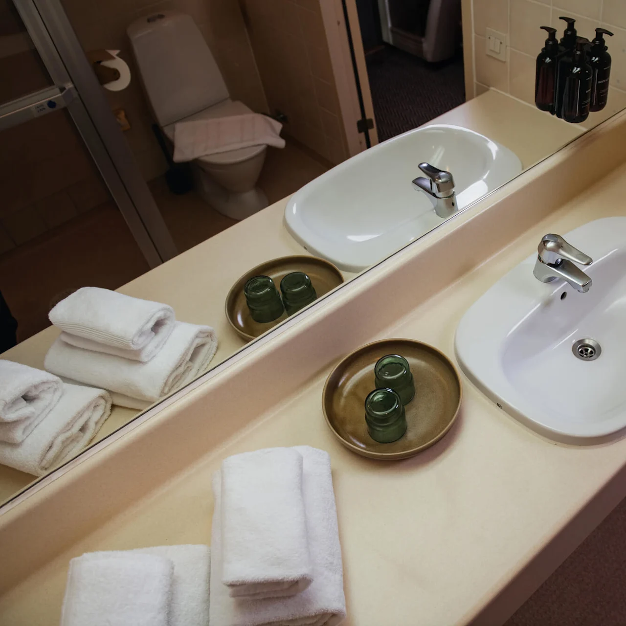 A white sink reflects on a mirror above a beige countertop. Rolled towels and small glass containers sit nearby. A toilet is visible in the background.