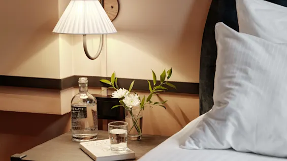 Bedside table holds a lamp, a bottle of water, a glass, a vase with white flowers, and a book. Adjacent to a neatly made bed with white linens.