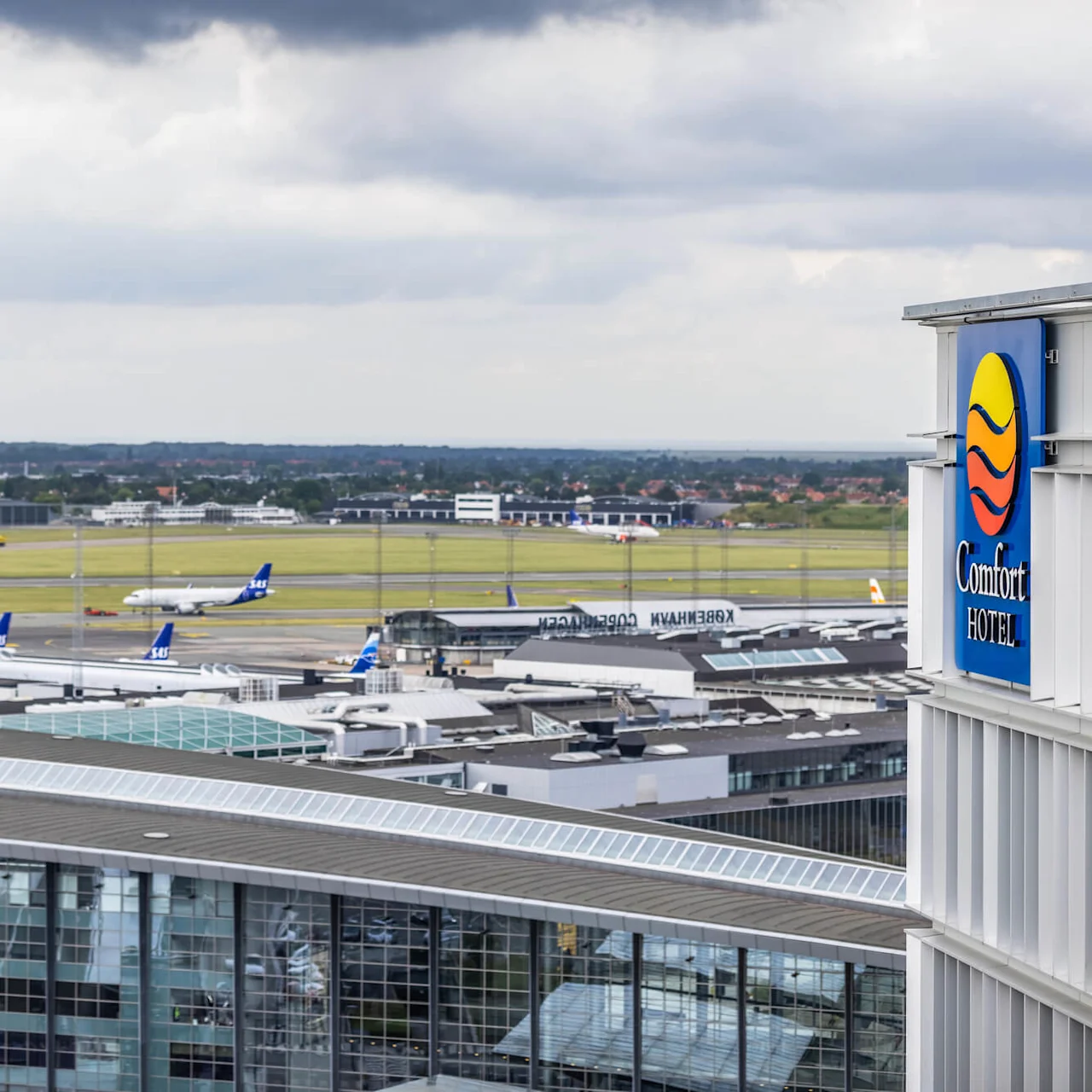 Fly på parkerte ved flyplassens gater, sett ovenfra, med moderne terminalbygg. Et "Comfort Hotel"-skilt er synlig, og det fjerne landskapet strekker seg under en skyete himmel. Tekst: "København."