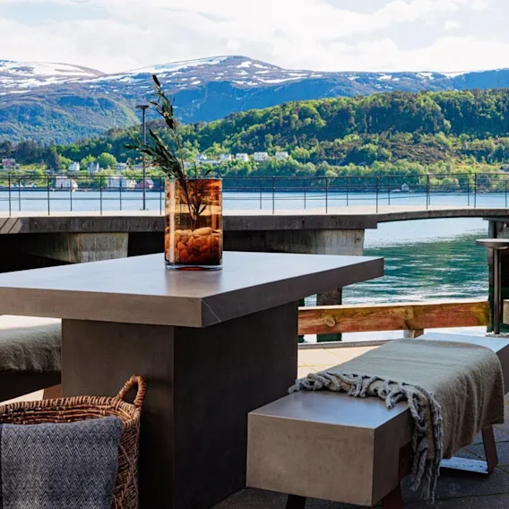 Et moderne utendørs bord med benker er utstyrt med koselige tepper, en plante i en glassvase på toppen, med utsikt over en naturskjønn fjell- og innsjøutsikt under en klar himmel.