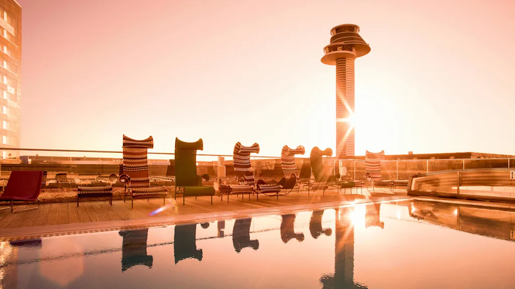 Poolstolar vänder mot en reflekterande pool vid soluppgången, bredvid ett modernt torn på en takterrass, med mjukt orange ljus.