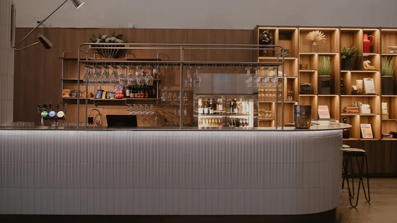 A sleek bar counter holds glasses and bottles, illuminated warmly; a built-in shelf displays plants and items, set within an elegant wooden theme.
