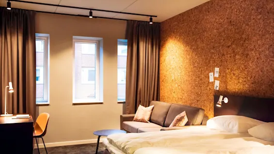 Bed with white linens sits against a cork wall, next to a gray couch in a warmly lit room. Three small windows reveal a brick building outside.