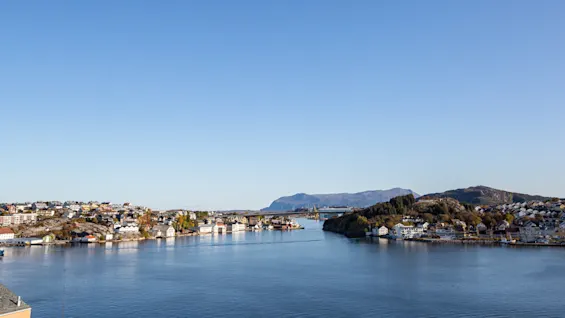 En bred elv strømmer mellom to befolkede bredder, hver med hus og trær, under en klar blå himmel med fjerne fjell.