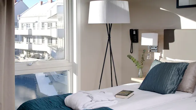 En pent oppredd hotellseng med turkise puter og en bok står ved et stort vindu. En gulvlampe og en telefon er i nærheten, med moderne bygninger utenfor.