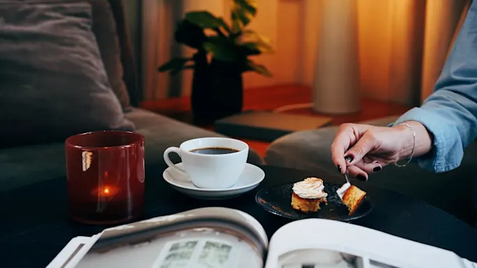 En person skjærer et stykke kake på en tallerken ved siden av en kaffekopp, med en tent lampe og plante i bakgrunnen.