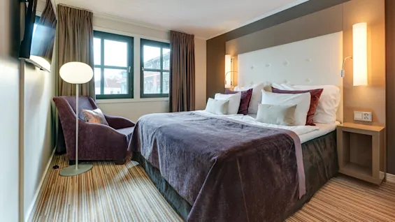 En säng med lila sängkläder står med nattduksbord i ett hotellrum. En vadderad stol och golvlampa står nära fönstret, bredvid den monterade TV:n.