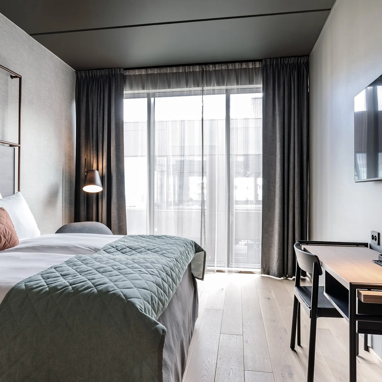 En prydligt bäddad säng med texturerade kuddar och vadderat täcke står i ett modernt hotellrum, bredvid en väggmonterad TV och ett skrivbord med en lampa och kaffekoppar.
