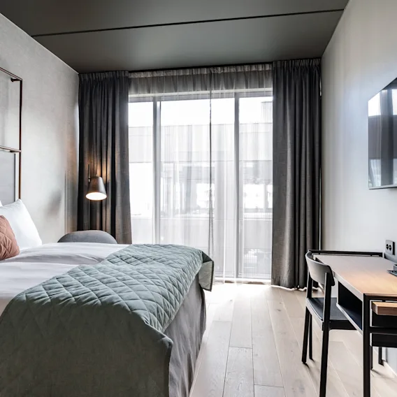 En pent oppredd seng med teksturerte puter og quiltet teppe er i et moderne hotellrom, ved siden av en veggmontert TV og et skrivebord med en lampe og kaffekopper.