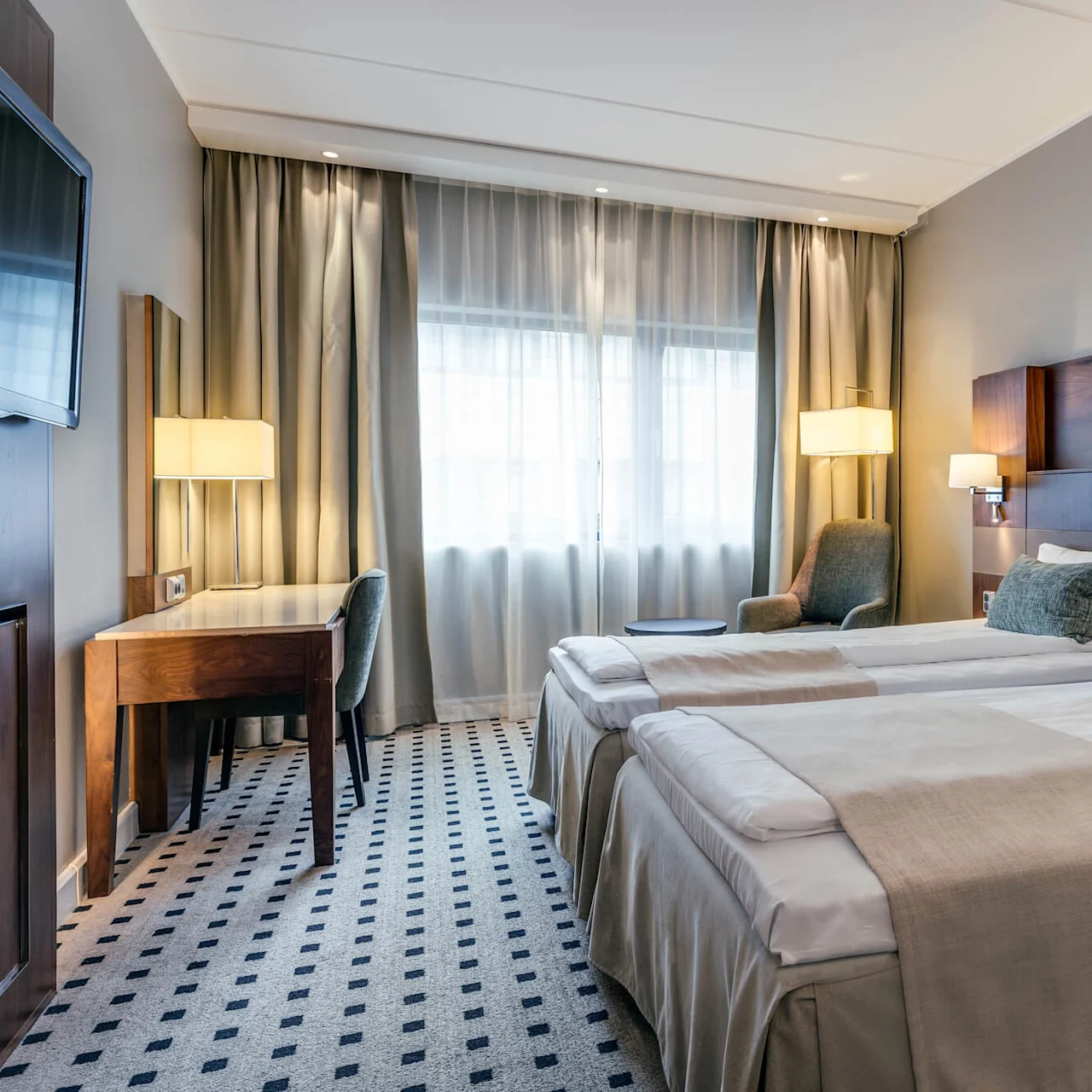 Twin beds with gray pillows and beige throws, positioned in a modern hotel room. There’s a desk with a chair and lamp beside a large window with curtains.