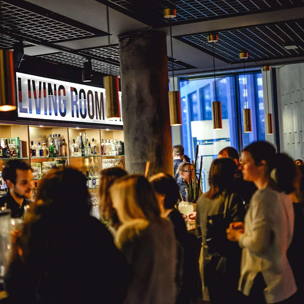 Människor umgås i en livlig bar med en bartender som serverar drinkar under en skylt med texten "LIVING ROOM".