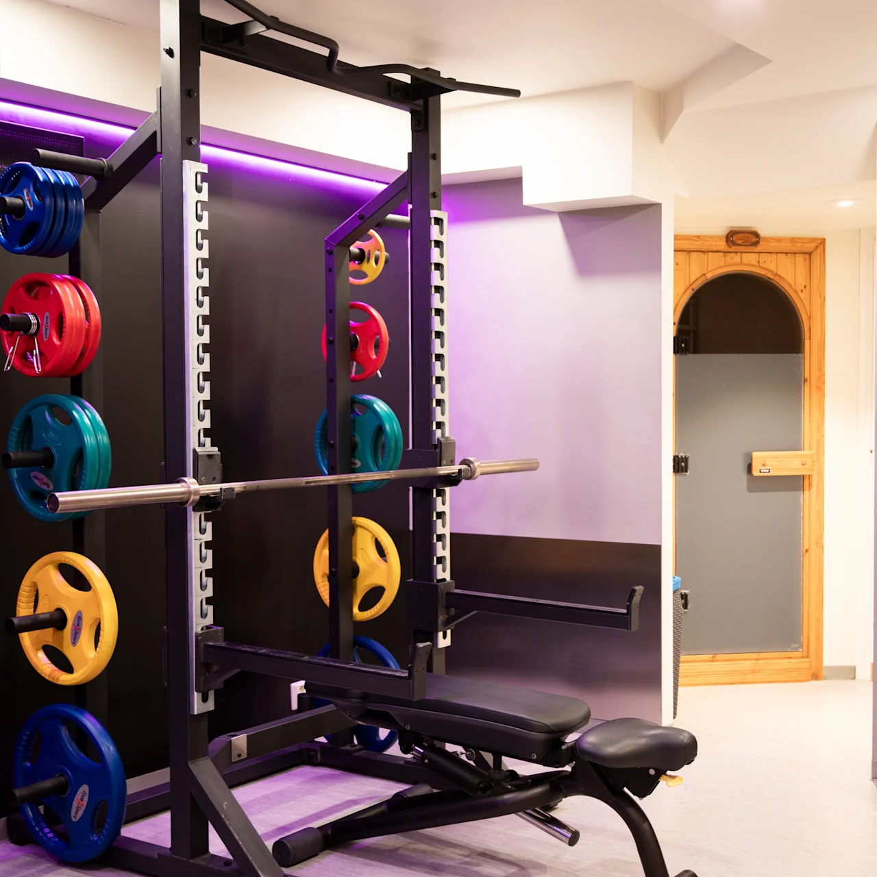 Vægtstang hviler på et squat-stativ med farverige vægtskiver; placeret i et fitnesscenter med lilla belyst væg, nær en dør og ved siden af en vandkøler og plante.