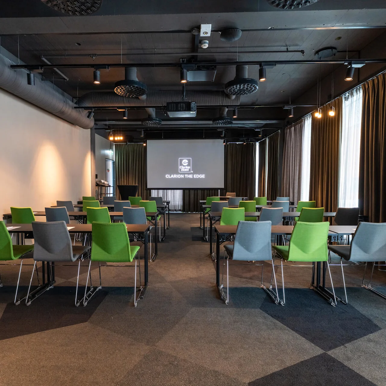 Konferencelokale med grønne og grå stole, der vender mod en skærm med logoet "Clarion the Edge"; mørke loftkanaler og gardindækkede vinduer i baggrunden.