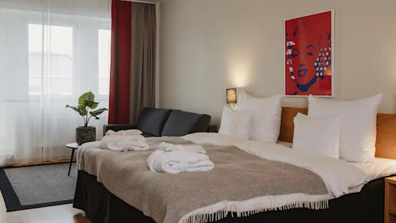 A neatly made bed with two robes lies in a cozy hotel room. A small couch and table with a plant are by the window. A colorful portrait adorns the wall.