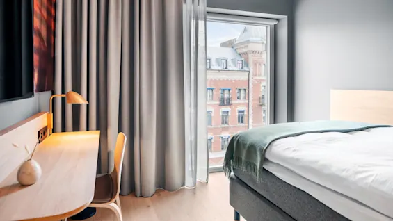 Et pent arrangert hotellrom med en seng med et grønt teppe, et skrivebord i tre med en lampe og stol, og et stort vindu som viser en teglbygning utenfor.