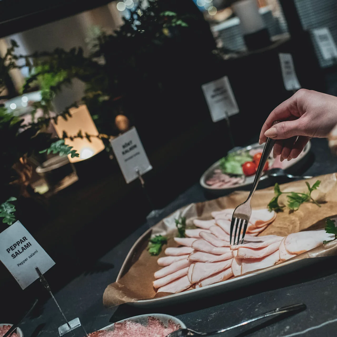 A hand uses a fork to pick up sliced meat from a platter labeled "PEPPAR SALAMI" in a buffet setting, surrounded by greenery and other dishes.