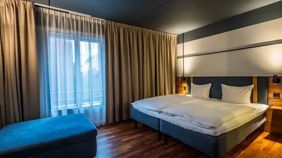 A neatly made bed with white linens stands against a wall in a cozy room, illuminated by soft lighting. Curtains cover a large window, paired with a blue ottoman. 