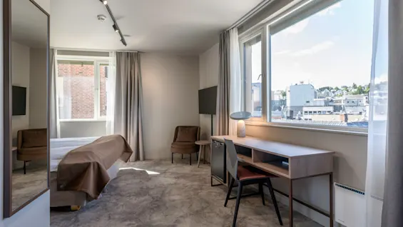A desk with a chair is positioned near large windows showing an urban view, while a bed with brown bedding is partially visible, surrounded by modern decor and neutral colors.