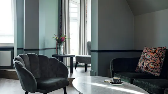 A velvet chair rests beside a sleek marble table holding a coffee cup in a softly lit room with large windows draped in curtains. A flower vase sits on a side table.