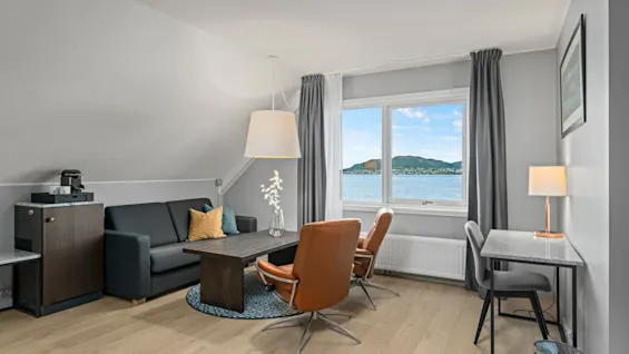 Et opholdsrum har en grå sofa og to orange stole omkring et træbord, med udsigt over havet gennem et stort vindue. Neutrale toner skaber en rolig atmosfære.