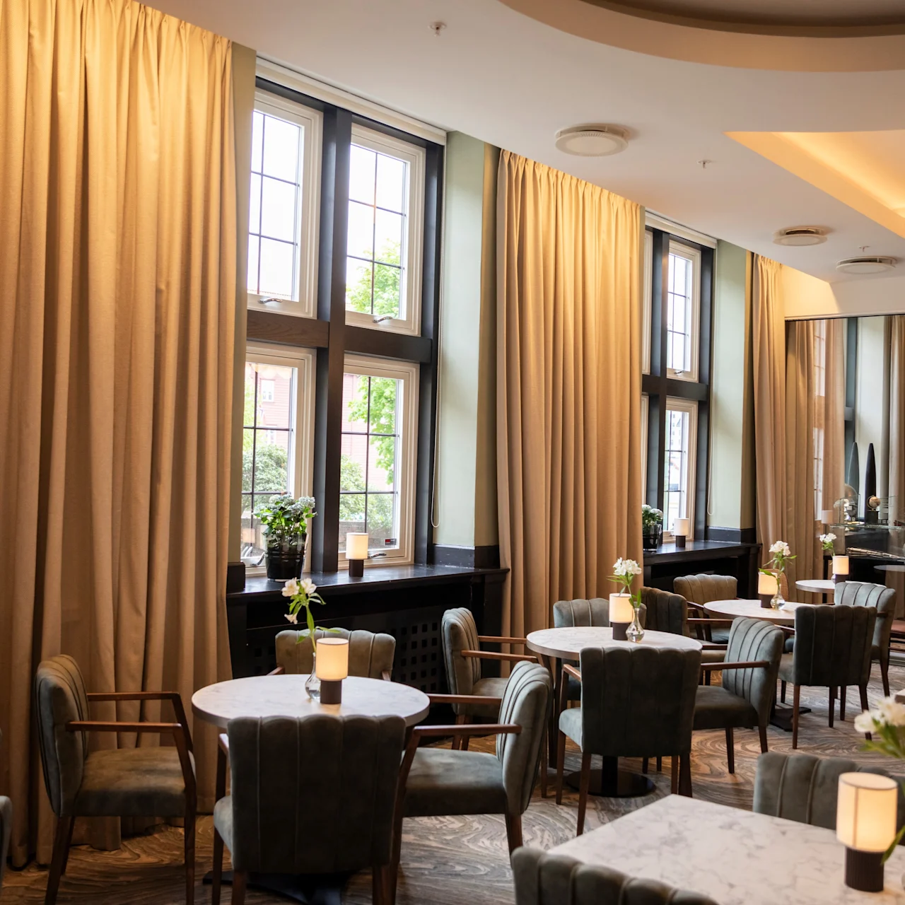 Round tables surrounded by cushioned chairs are arranged in a bright room with large windows and beige curtains, set for dining with vases and candles on tables.