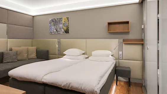 En snyggt bäddad dubbelsäng står bredvid en grå soffa i ett modernt hotellrum, med en vägghylla och abstrakt konst ovanför. Mjuk belysning förstärker den mysiga atmosfären.