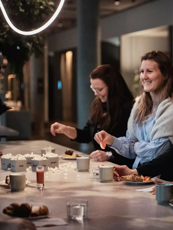 Fyra kvinnor ler och umgås vid ett bord med drycker och snacks i ett svagt upplyst rum.