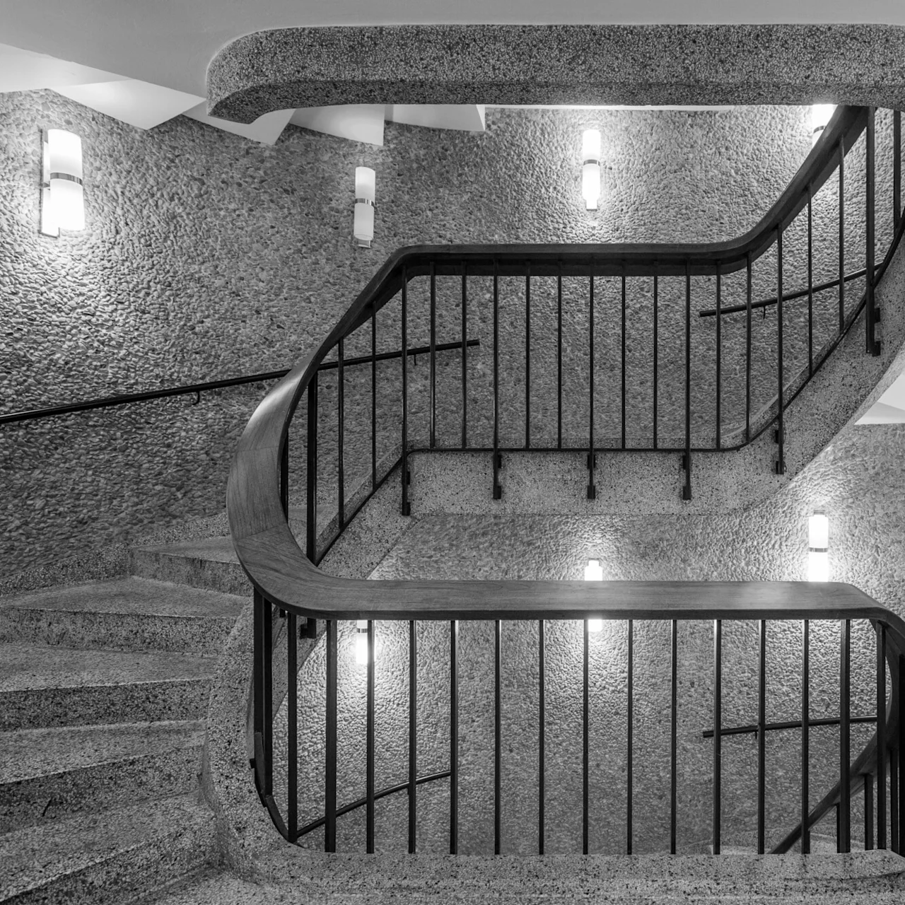 Curved staircase with wrought iron railing ascends between textured stone walls, illuminated by wall-mounted lights, creating a modern architectural aesthetic.