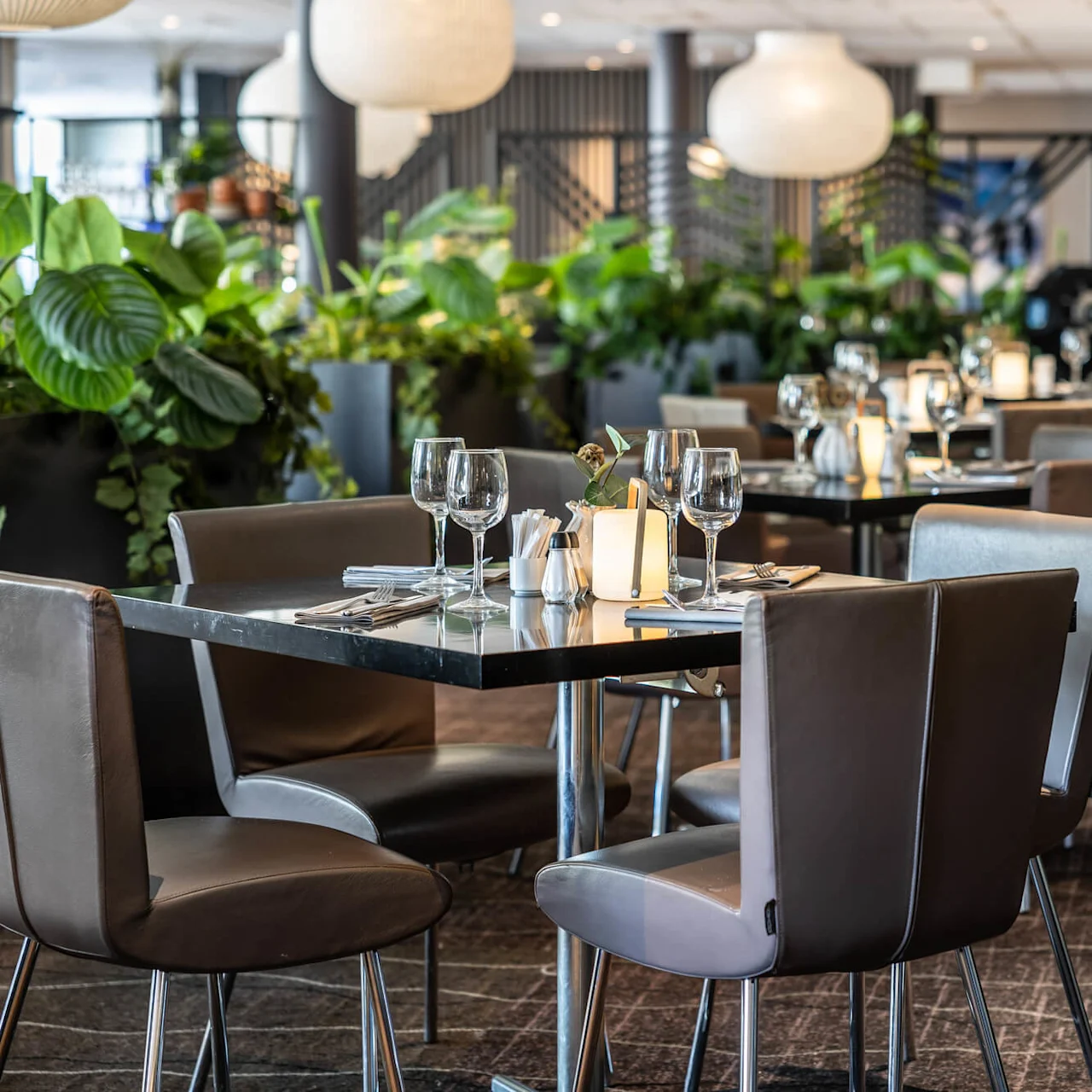 Elegante spiseborde dækket med glas og servietter omgivet af komfortable stole. Indendørs planter og moderne belysning skaber en frodig, indbydende restaurantatmosfære.