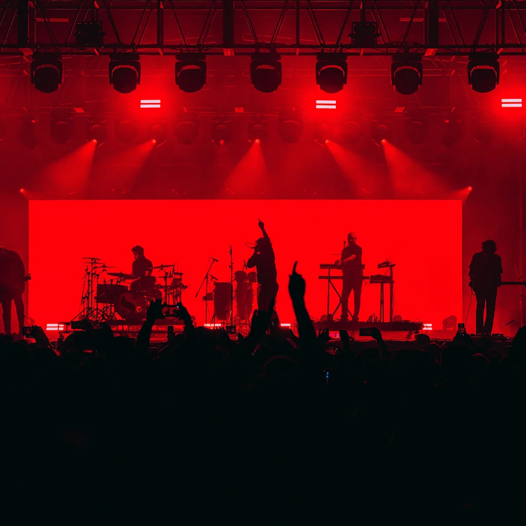 Ett band uppträder på en scen badad i rött ljus, med en stor publik silhuetterad i förgrunden. Två dubbelstreckssymboler syns.