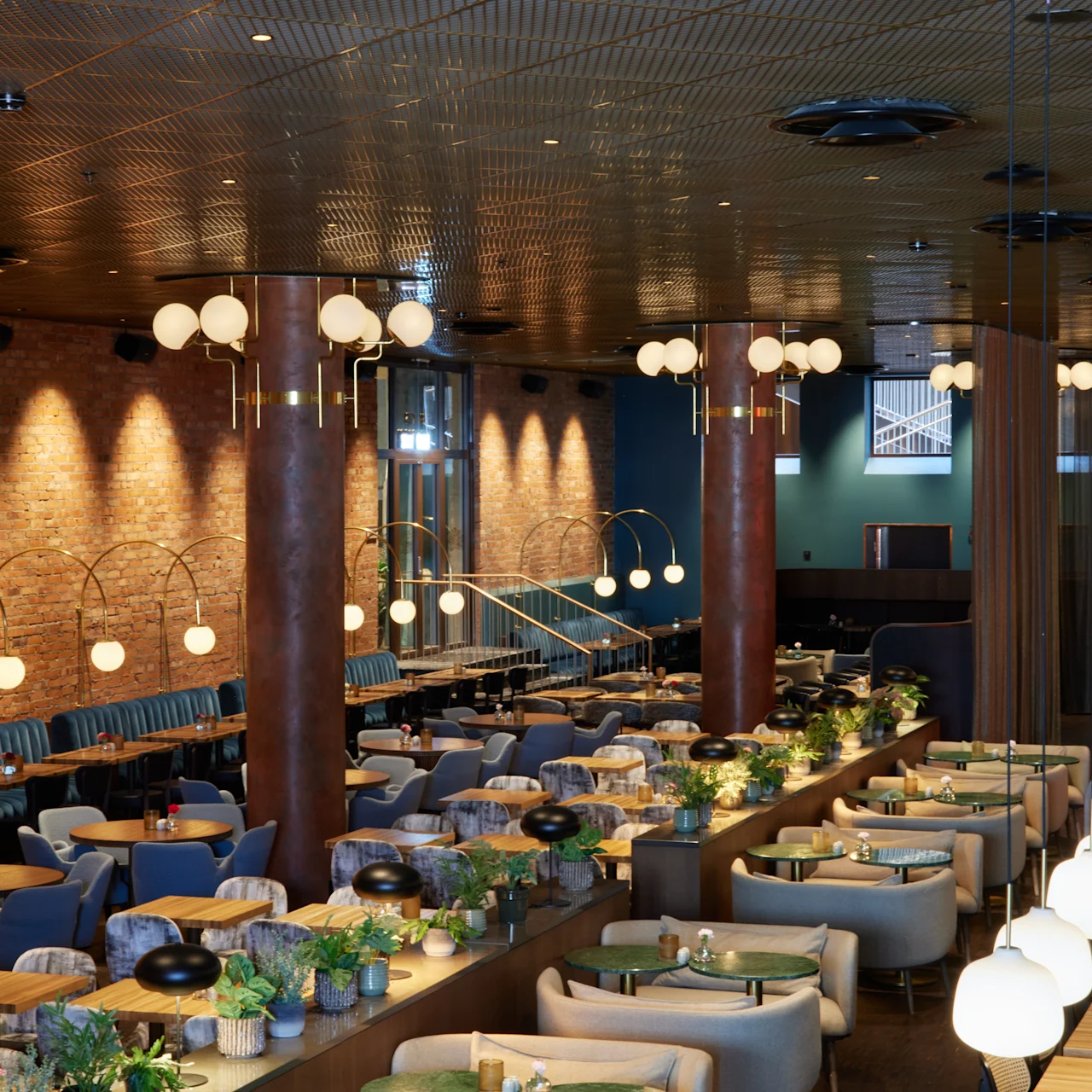 Snygg restauranginteriör med eleganta matbord dekorerade med växter, omgivna av bekväma sittplatser. Varm belysning från sfäriska lampor betonar tegelväggarna, vilket skapar en mysig, elegant atmosfär.