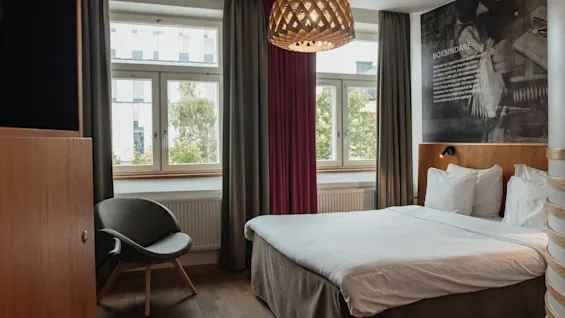 Home Hotel Uppsala: A modern room with a comfortable bed, stylish chair, large window, and a wall mural featuring BOKBINDARE.
