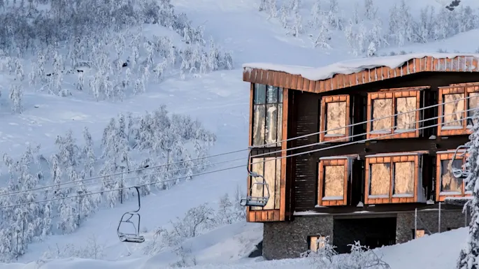 A wooden building with large windows, covered in snow, stands on a snowy mountainside. Nearby, a ski lift ascends towards snow-laden trees under a clear sky.