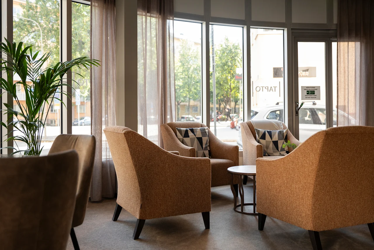 Bekväm sittgrupp på Home Hotel Tapto med fåtöljer, en växt och stora fönster mot en gata. Text: TAPTO