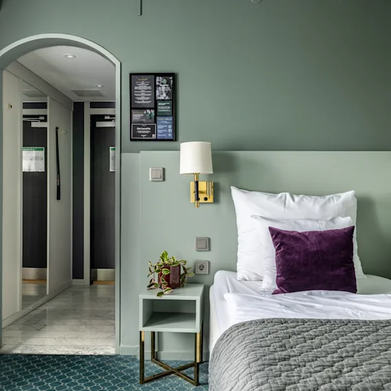 Ett mysigt hotellrum med en enkelsäng, vita och lila kuddar. Mjuka gröna toner skapar en lugnande atmosfär, komplementerat av en växt på ett bord. En korridor i bakgrunden ger djup.