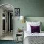 Kodikas hotellihuone, jossa on yksi vuode koristeltuna valkoisilla ja violetilla tyynyillä. Pehmeät vihreät sävyt luovat rauhoittavan ilmapiirin, jota täydentää pieni kasvi vieressä. Taustalla käytävä tuo syvyyttä.
