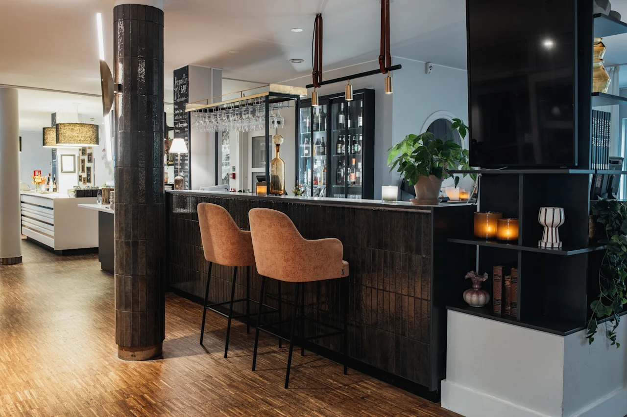 En hyggelig hotelbar med to tomme stole, en velassorteret disk og varm belysning, klar til gæster.