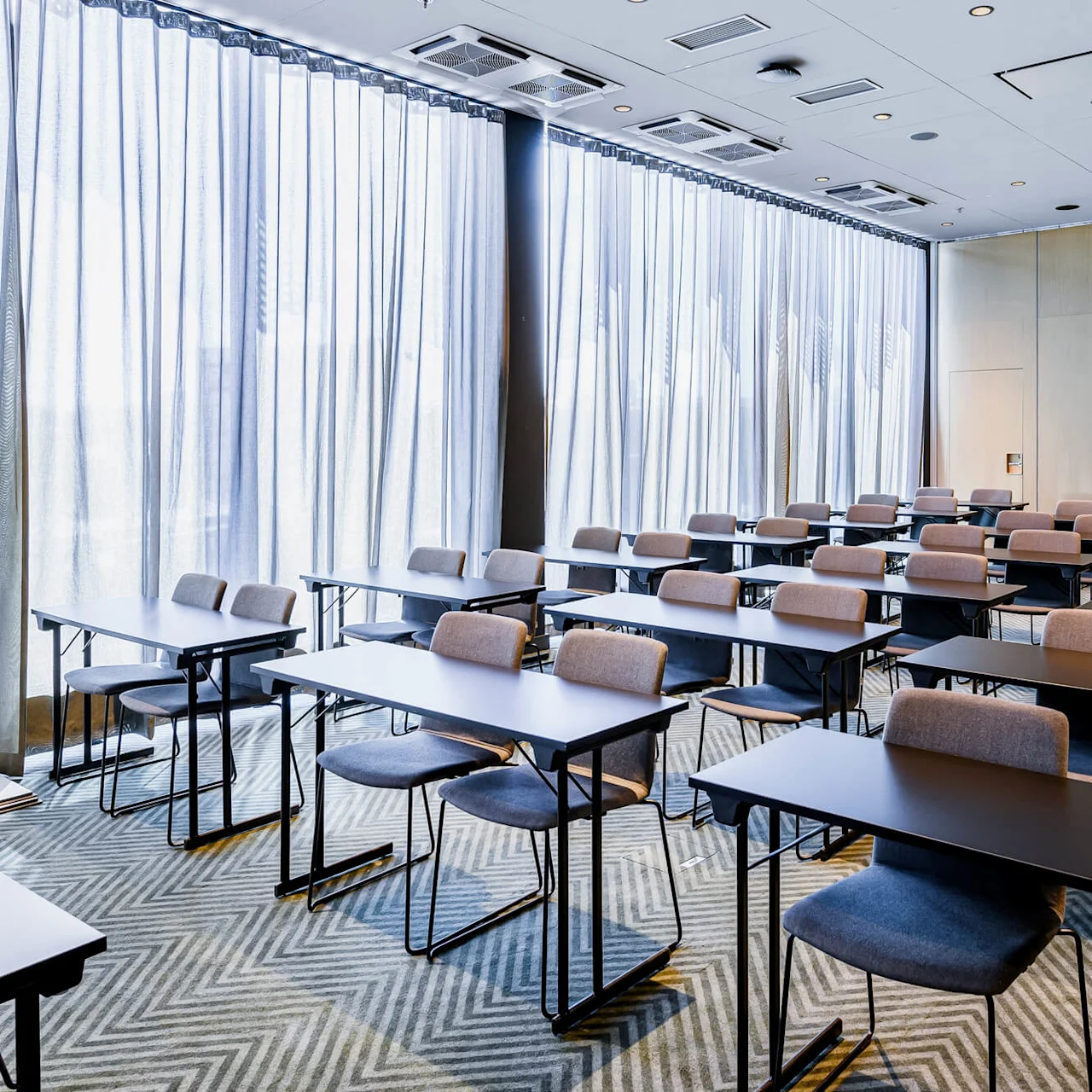 Rows of empty tables and chairs organized in a classroom setup, facing a wooden wall. Soft natural light filters through large, sheer curtained windows on the left side.