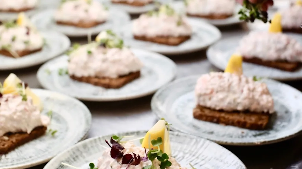 Norwegian bokmål: Smørbrød med kremet topping og sitronkile på tallerkener. Hånd pynter med urter på et kjøkken.