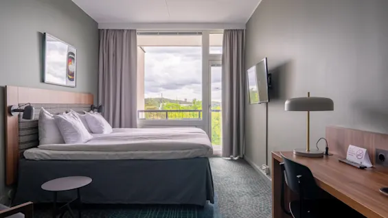 En pent og ryddig dobbeltseng med hvitt sengetøy står i et hotellrom. Et vindu viser utsikt over grøntområde. Et skrivebord med stol og lampe er i nærheten.