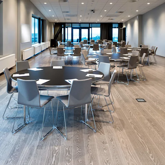 Konferanserom med runde bord og grå stoler ordnet pent, med store vinduer. Papirer og penner på bordene. Taket har rutenettmønster med lys og projektorer.