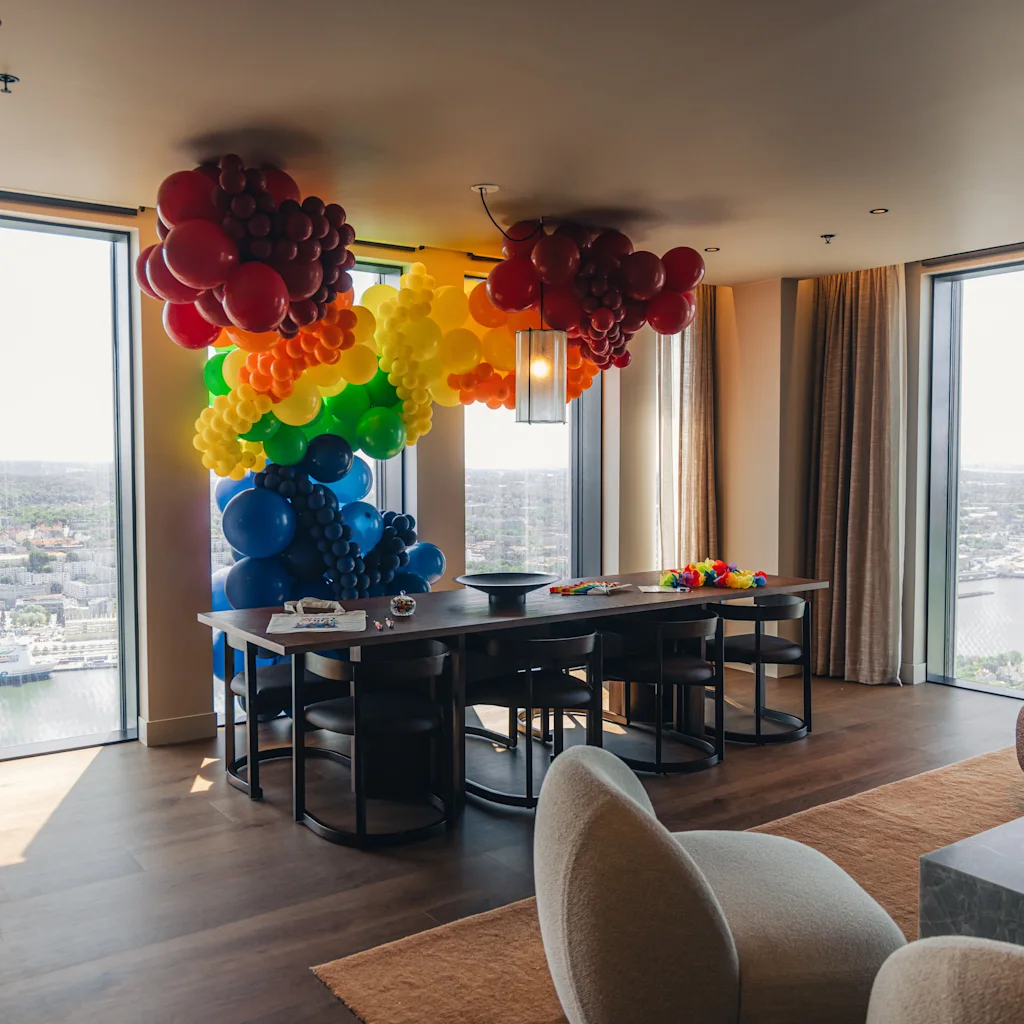 Colorful balloon arch stretches over a dining table in a modern apartment with wooden floors, large windows, and city views. Interior features couches and rainbow flags for decoration.