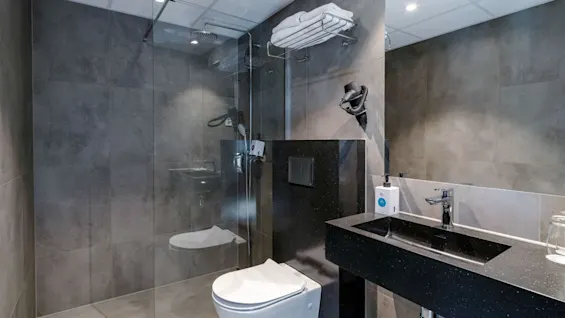 Moderne badeværelse med sort vask, væghængt toilet og indglasset brusebad. Hårtørrer og håndklæder er monteret på væggen. Grå fliser dækker gulv og vægge.