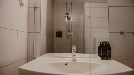 Sink reflecting in a bathroom mirror, flanked by toiletry bottles, facing a shower with wall-mounted soap dispensers, enclosed in sleek, tiled walls.