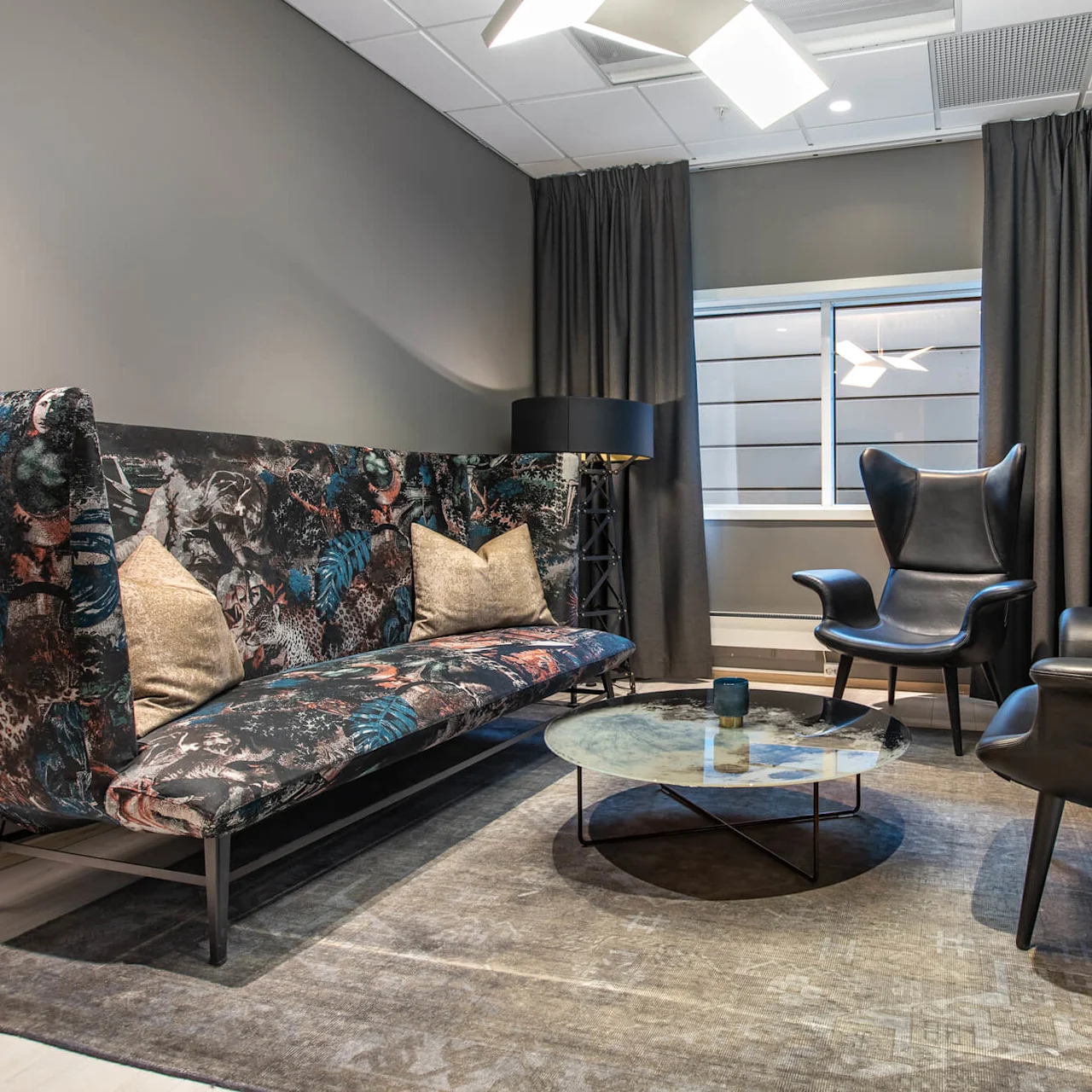 Colorful sofa with patterned upholstery and plush pillows faces two black leather armchairs around a glass coffee table in a modern, elegantly lit room with large windows and curtains.