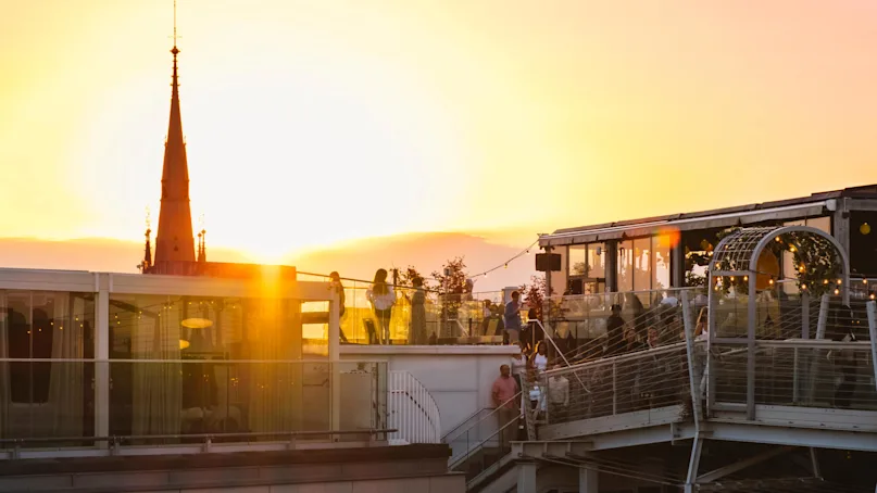 People gather on a rooftop terrace at sunset, with a church spire silhouetted in the background and string lights creating a warm ambiance.