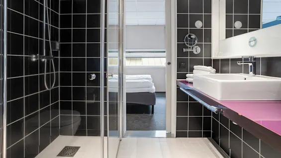 A modern bathroom with a glass-door shower, sink, and towels, visible through to a tidy bedroom with a made bed and a skylight.